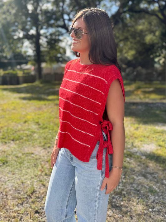 A woman wearing a red sleeveless knit top with thin white horizontal stripes and side ties, paired with light blue jeans, standing outdoors in a grassy park area with trees in the background.