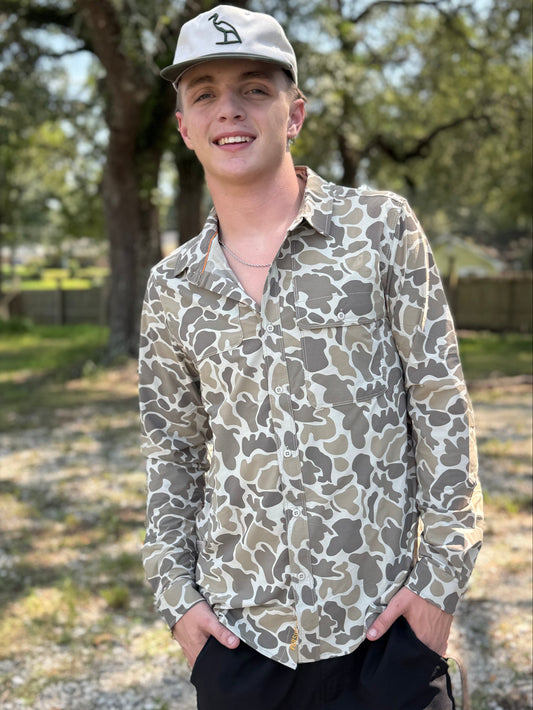 A young man standing outdoors in a wooded area, wearing a long-sleeve button-up shirt with an abstract camouflage pattern in olive green, brown, and beige, paired with black shorts and a baseball cap.