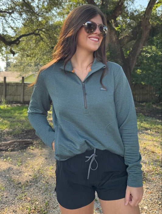 A woman wearing a ribbed slate green quarter-zip pullover and black shorts, standing outdoors in a wooded area with trees in the background.