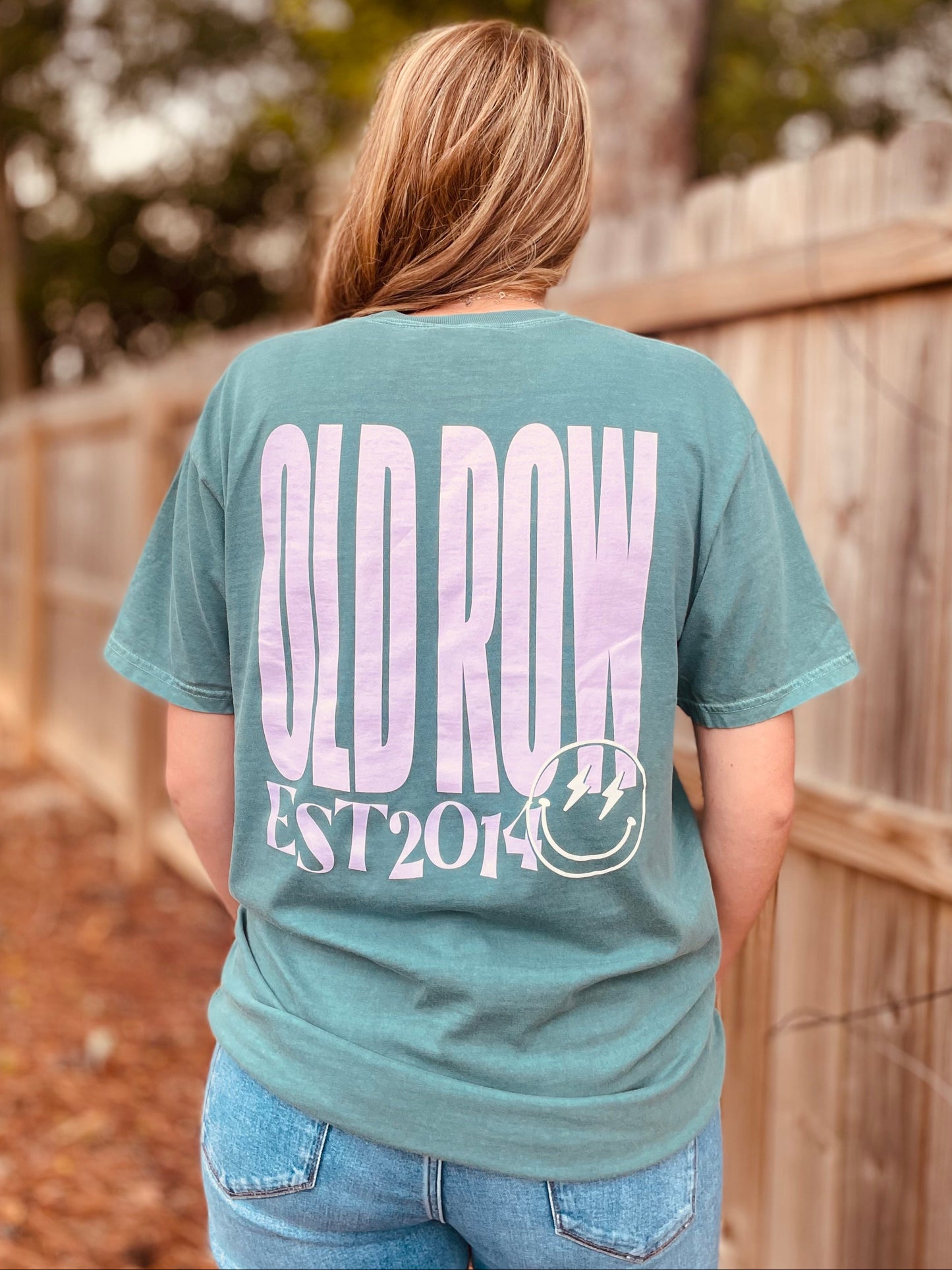 A woman viewed from behind wearing a faded teal graphic t-shirt with 'OLD ROW EST 2014' printed in light pink and white on the back, paired with blue jeans, standing outdoors in front of a wooden fence.