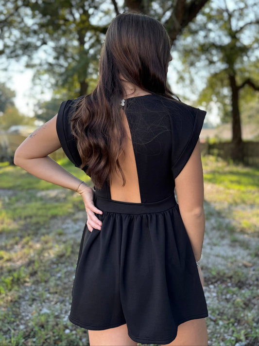 A woman viewed from behind wearing a black short-sleeve romper with a large open back, standing outdoors in a grassy area with trees in the background.