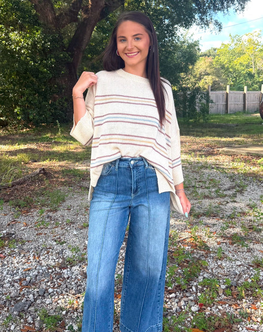 A woman standing outdoors in front of a large tree, wearing a cream-colored knit top with thin horizontal stripes, high-waisted wide-leg blue jeans, and brown flat sandals.