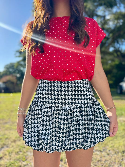 A woman outdoors wearing a bright red short-sleeve top with white polka dots and a black-and-white houndstooth skirt.