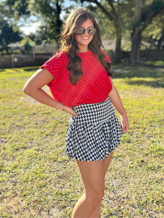 A woman wearing a red short-sleeve top with white polka dots and a black and white checkered mini skirt, standing outdoors on a grassy lawn with trees in the background.