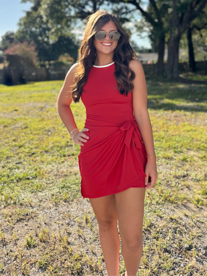 A woman wearing a sleeveless red wrap-style mini dress and sunglasses stands outdoors in a grassy park area with large trees in the background.