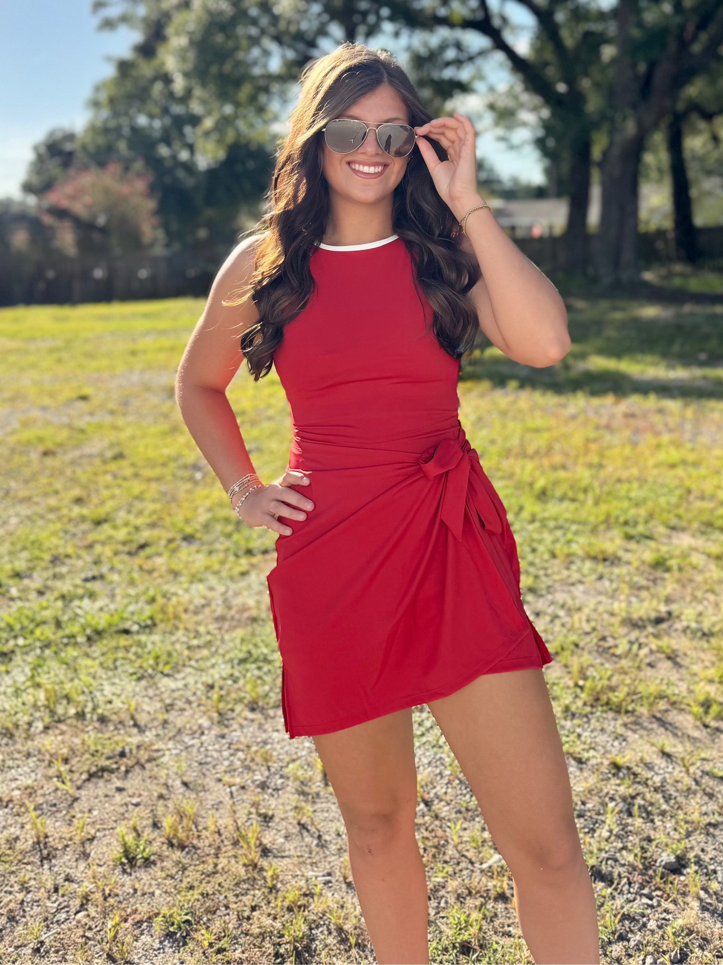 A woman wearing a sleeveless red mini dress with a side tie detail and sunglasses, standing in a grassy outdoor park with trees in the background.