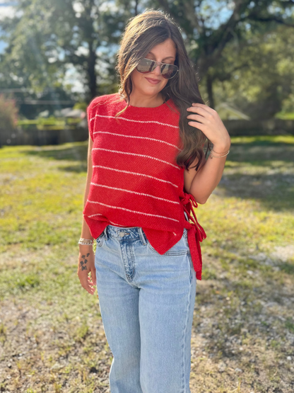 A woman wearing a red knit top with thin white horizontal stripes and side ties, paired with light-wash jeans, standing outdoors in a grassy park area with trees in the background.
